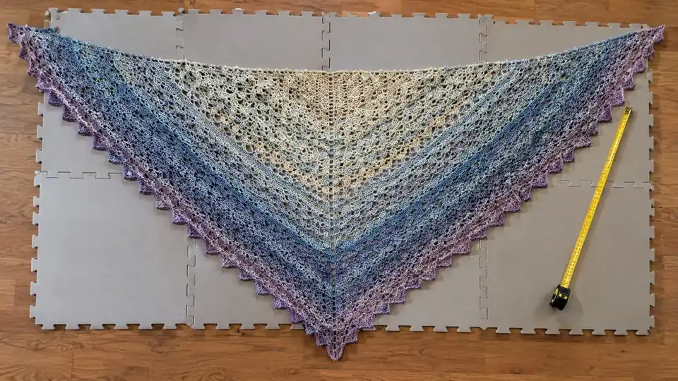 A crochet shawl laid out flat on blocking mats with a measuring tape beside it